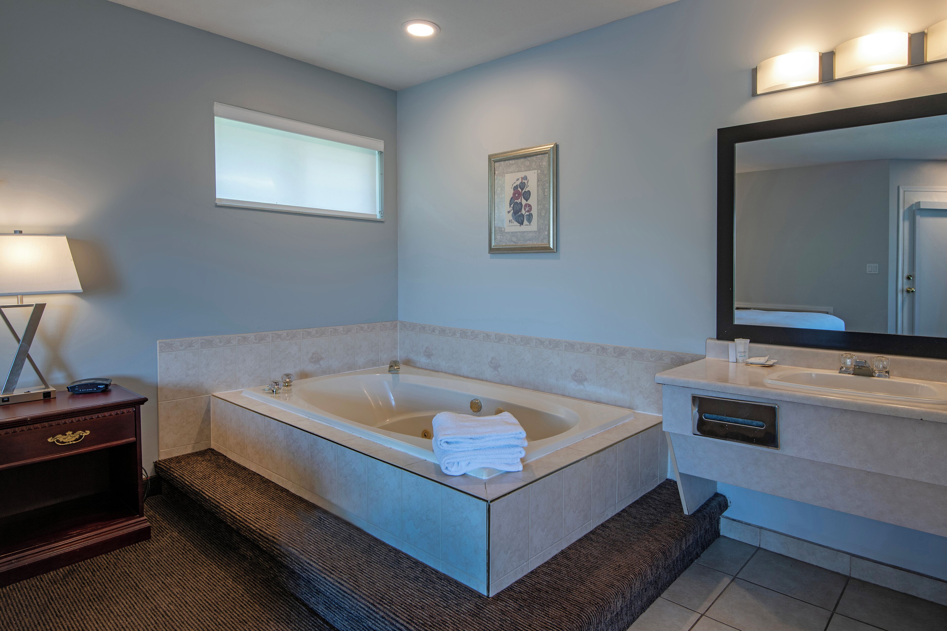 A modern bathroom with a large bathtub, a vanity with a mirror, and a window with blinds.