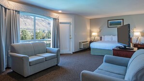 Habitación de hotel con cama, dos sillones, un escritorio con computadora, ventana con cortinas y un cuadro en la pared.