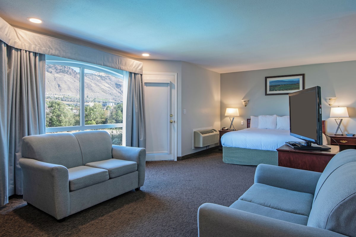 Une chambre d’hôtel comprenant un lit, deux fauteuils, un bureau avec un ordinateur, une fenêtre avec des rideaux et un tableau au mur.