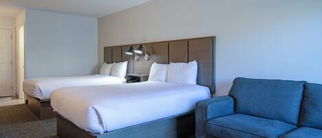 Habitación de hotel con dos camas, un sofá azul, una mesita pequeña y una lámpara fijada a la pared.