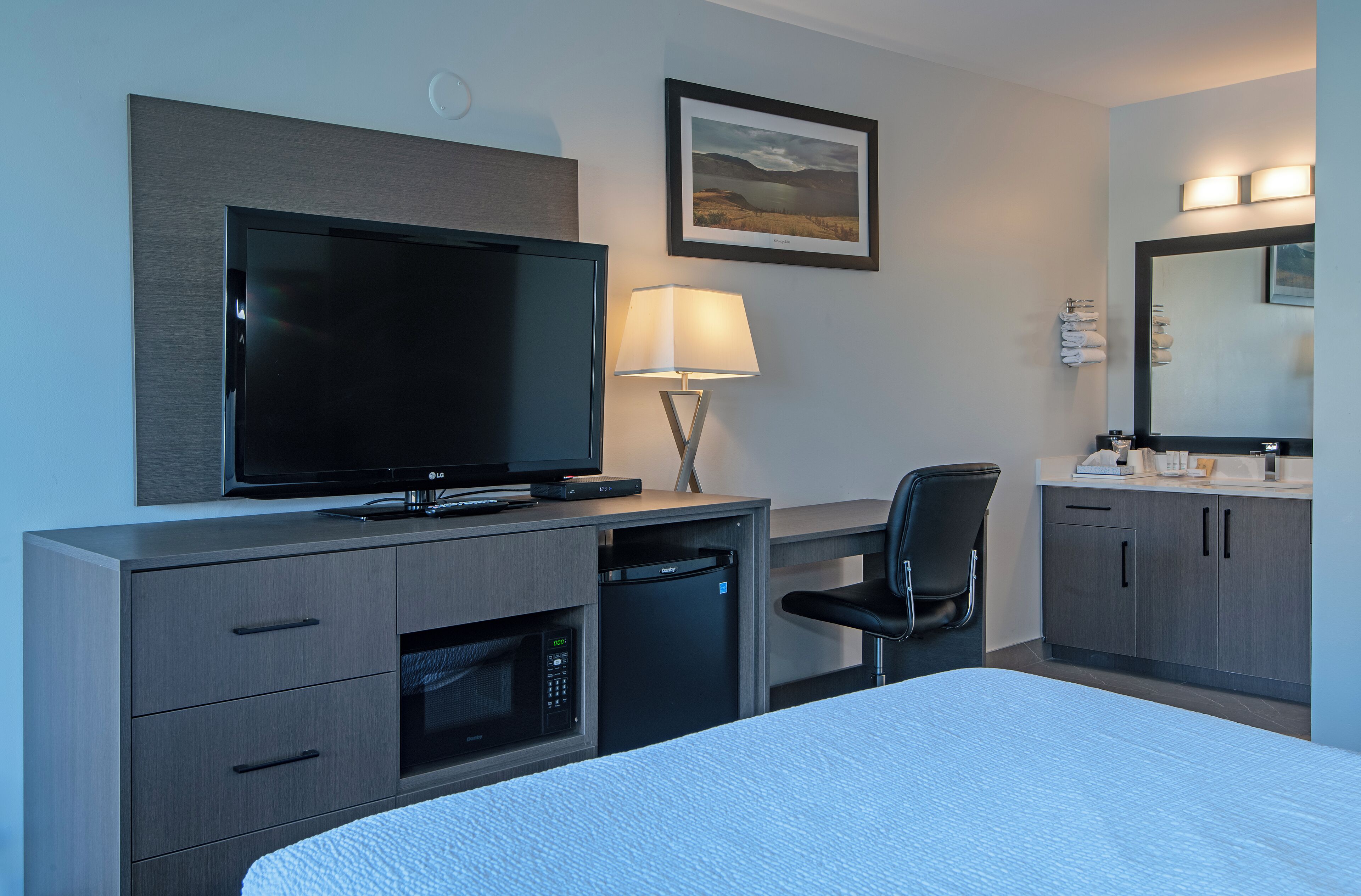 A hotel room with a bed, a TV, a desk with a lamp, a chair, a microwave, a mini fridge, a bathroom with a sink and mirror, and a framed picture on the wall.
