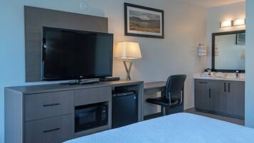 A hotel room with a bed, a TV, a desk with a lamp, a chair, a microwave, a mini fridge, a bathroom with a sink and mirror, and a framed picture on the wall.