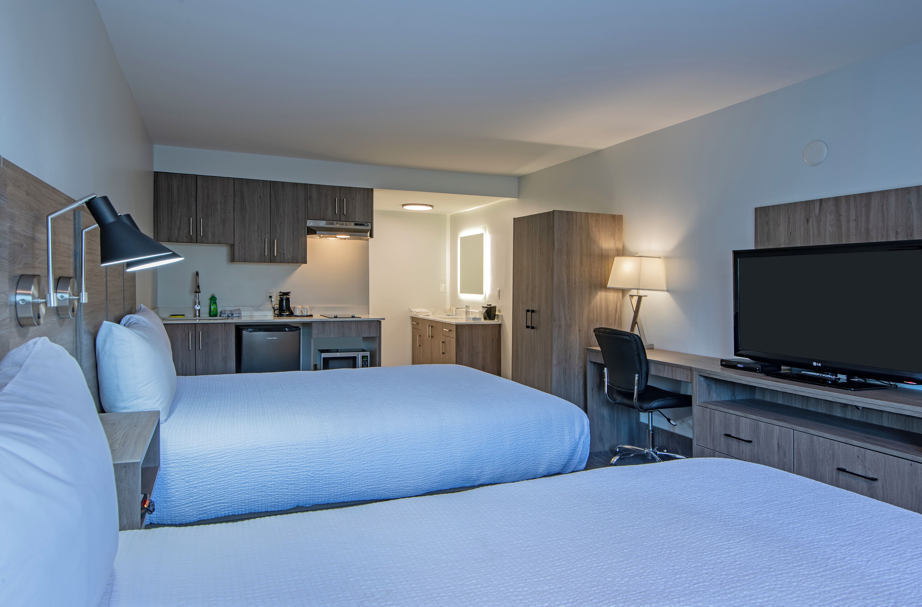 A hotel room with two beds, a desk, a TV, and a kitchenette.