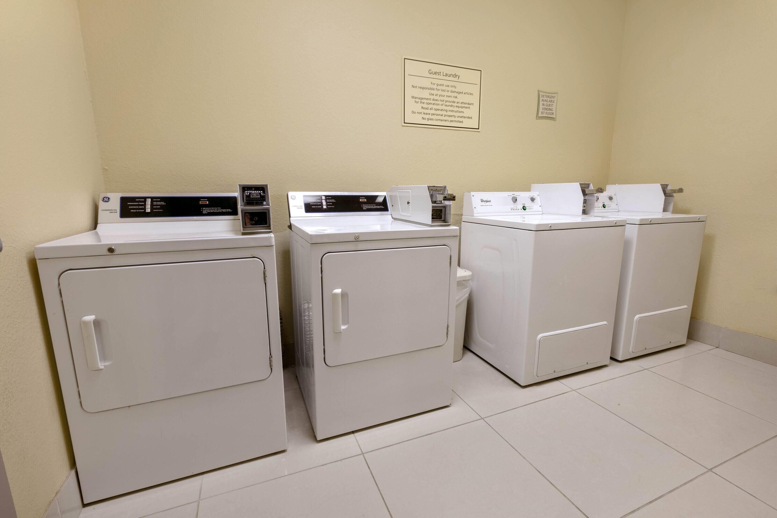 A laundry area with four white front-loading washers and dryers, a sign indicating guest laundry use, and a wall-mounted sign with instructions.