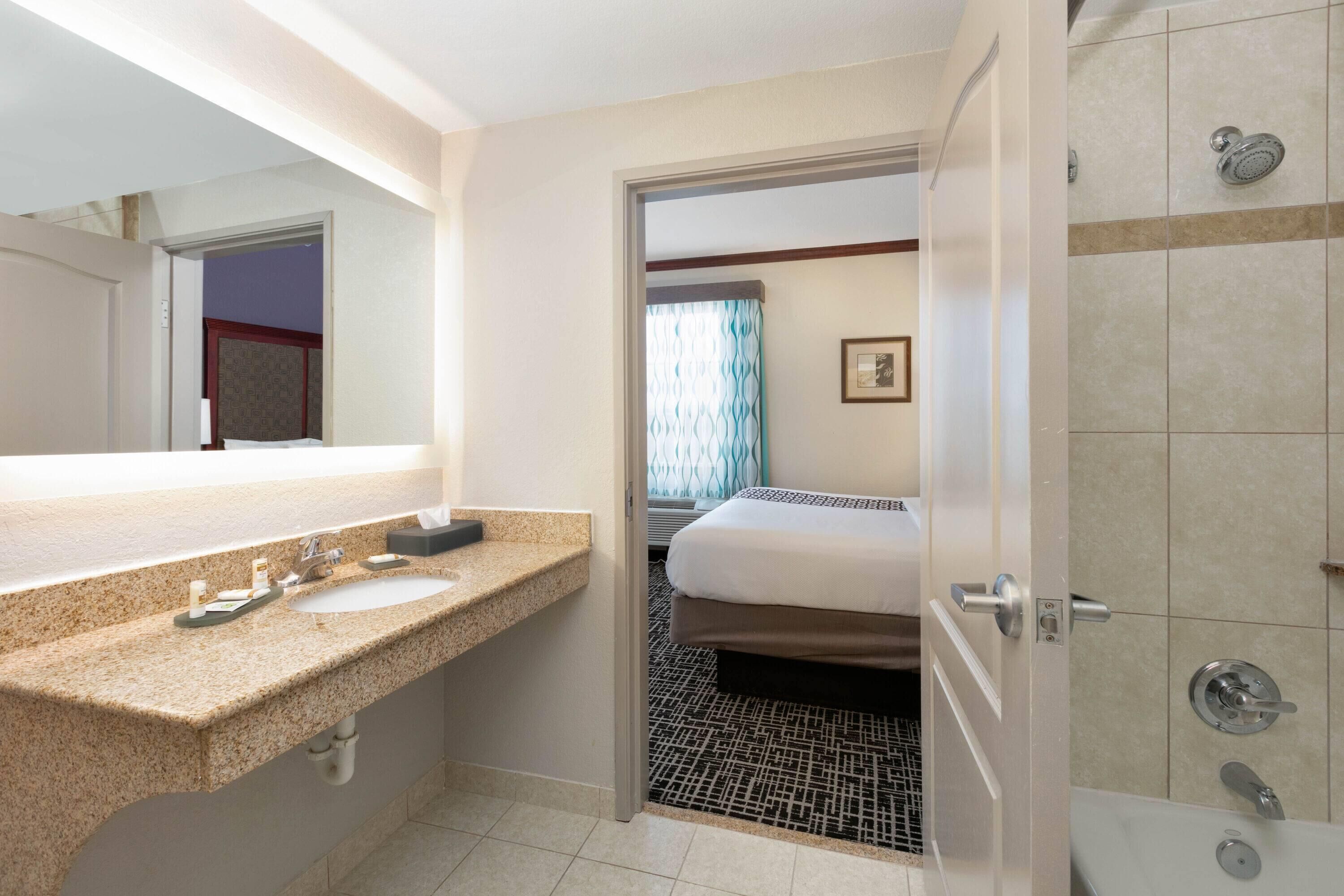 A hotel bathroom with a double sink, a shower, and a bed visible through an open door.
