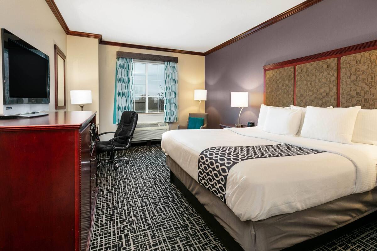 A hotel room with a large bed, a desk with a TV, two lamps, a chair, and a window with curtains.