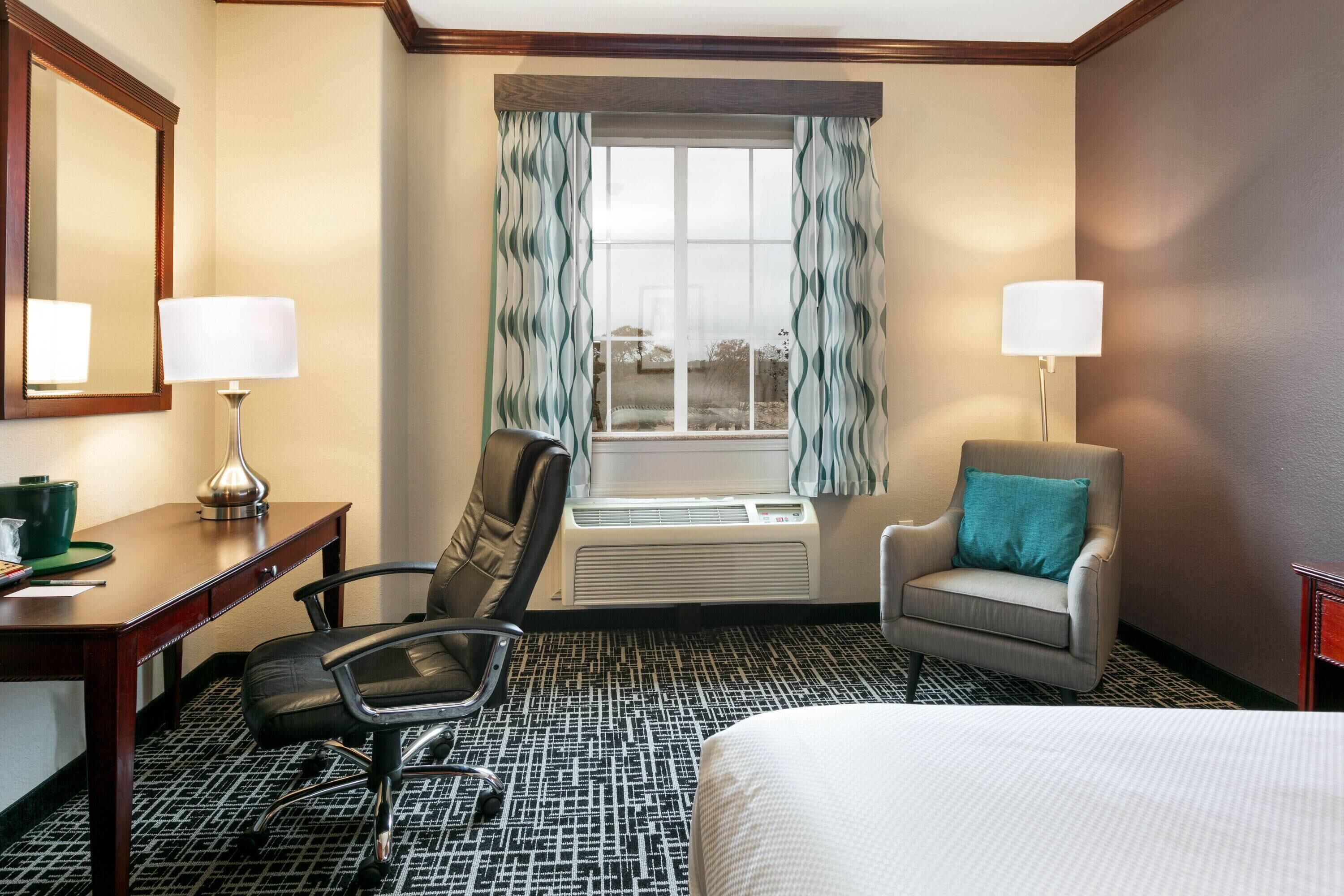 A hotel room with a bed, a desk with a lamp, a chair, a window with curtains, and an air conditioning unit.