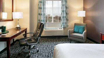 A hotel room with a bed, a desk with a lamp, a chair, a window with curtains, and an air conditioning unit.