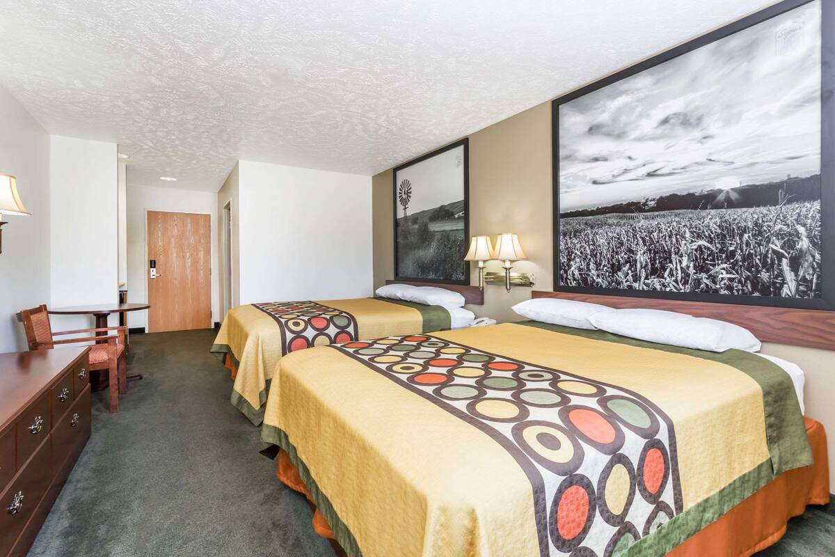 A hotel room with two beds, a desk, a chair, a wardrobe, and a large framed picture of a field.
