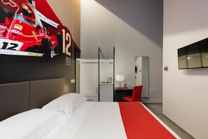 A hotel room with a large bed, a desk with a chair, a TV, and a wall with a racing car poster. - Hotel Maranello Village (Maranello)