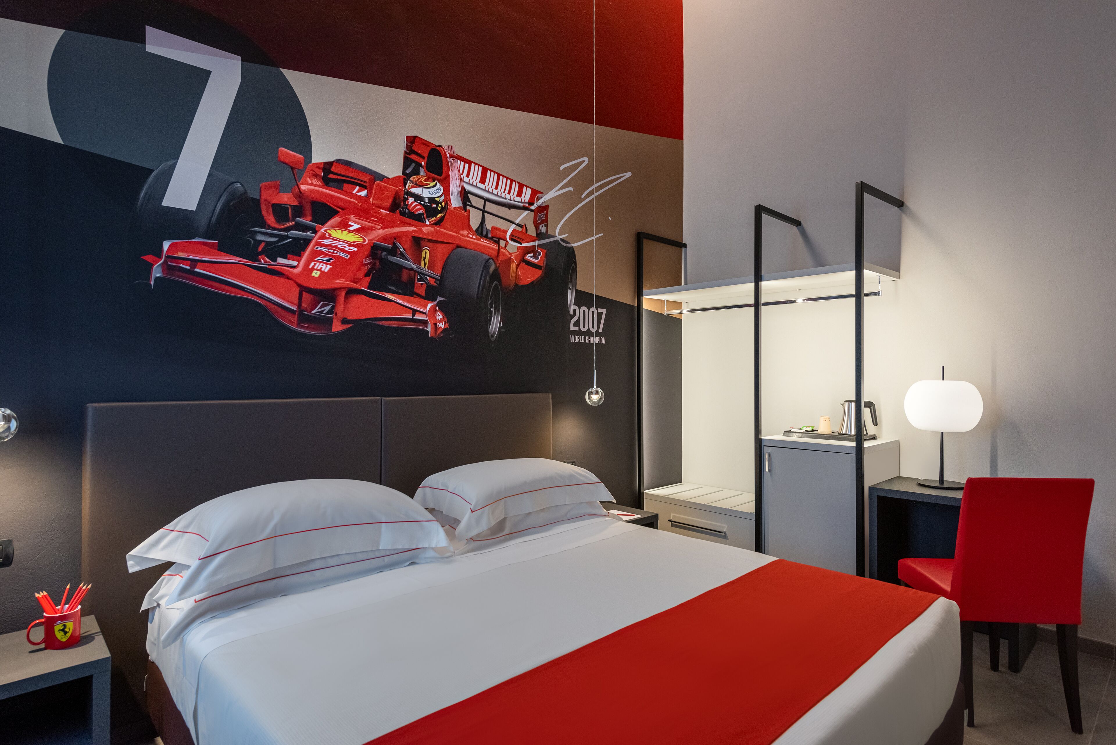 A hotel room with a bed, a desk, a chair, a nightstand, and a wall with a large number and a racing car illustration.