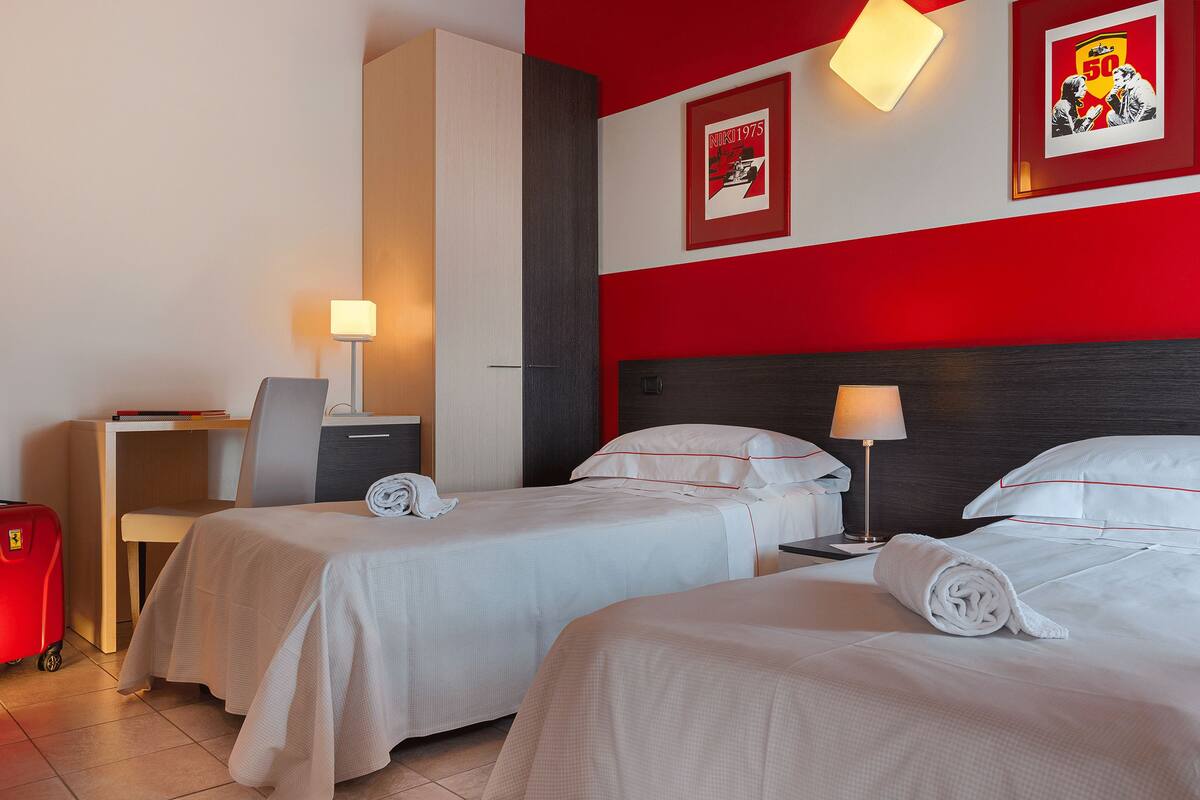 A hotel room with two beds, a desk, a chair, a red suitcase, and a wall with a framed picture.