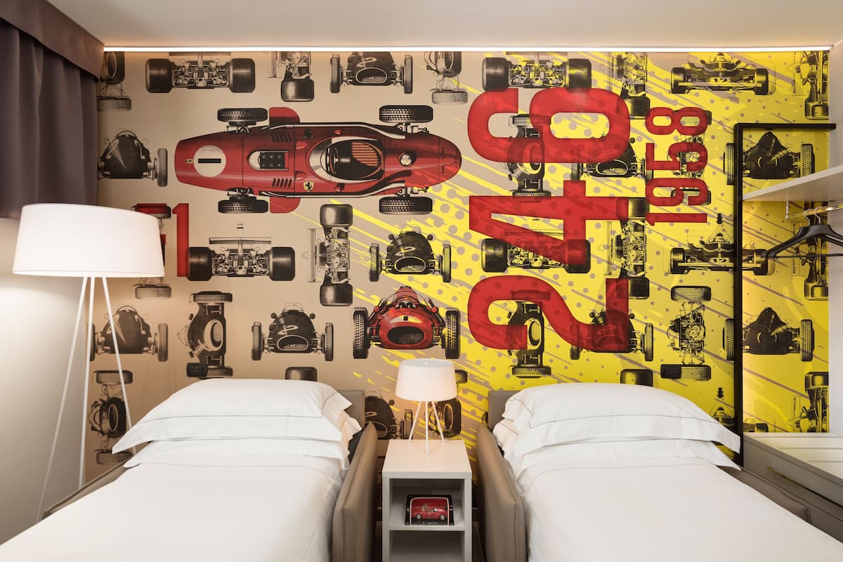 A hotel room with two beds, a nightstand with a lamp, and a wall decorated with racing car decals.