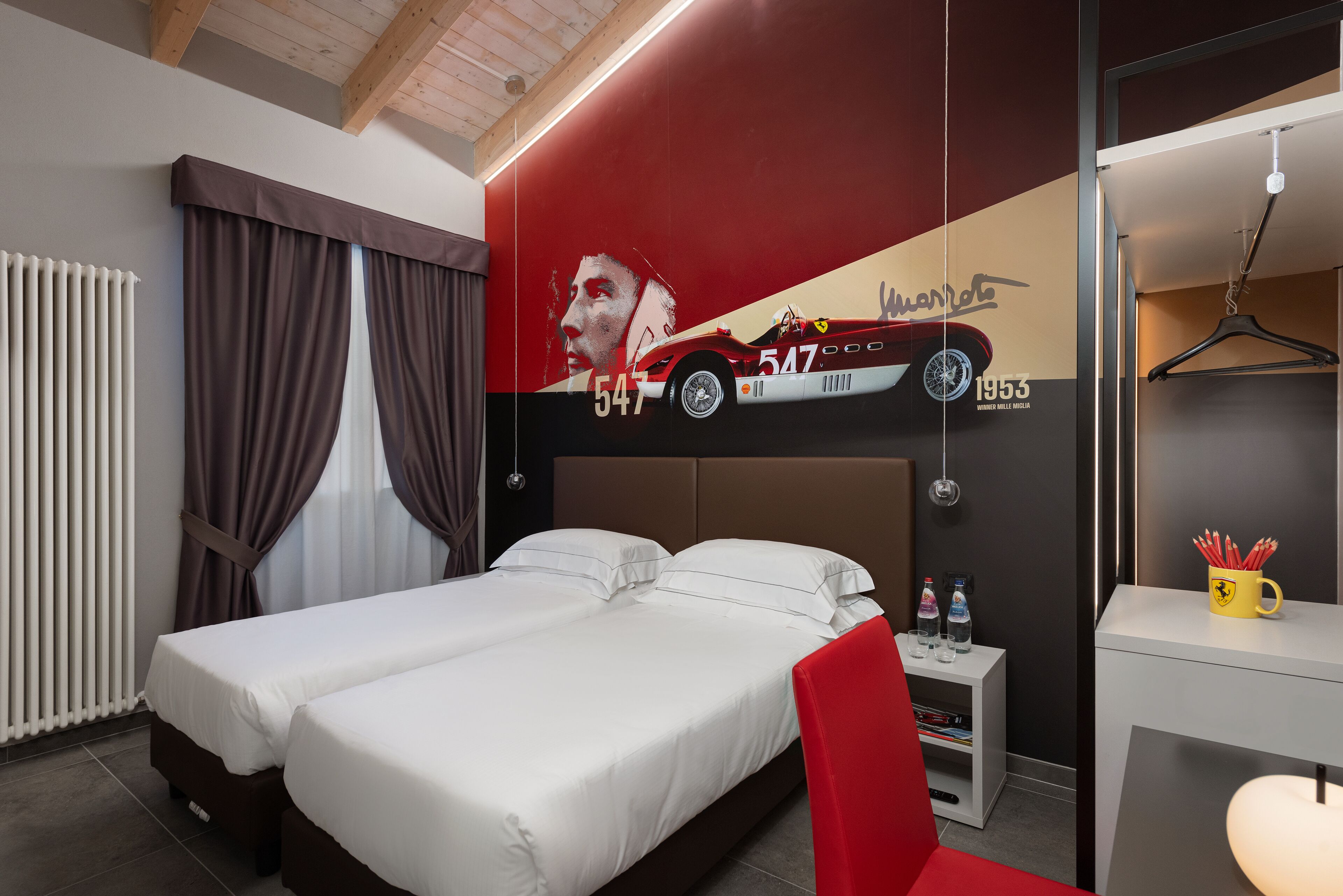 Quarto de hotel com uma cama, uma cadeira vermelha, uma pequena mesa com garrafas e uma parede com um carro e números.