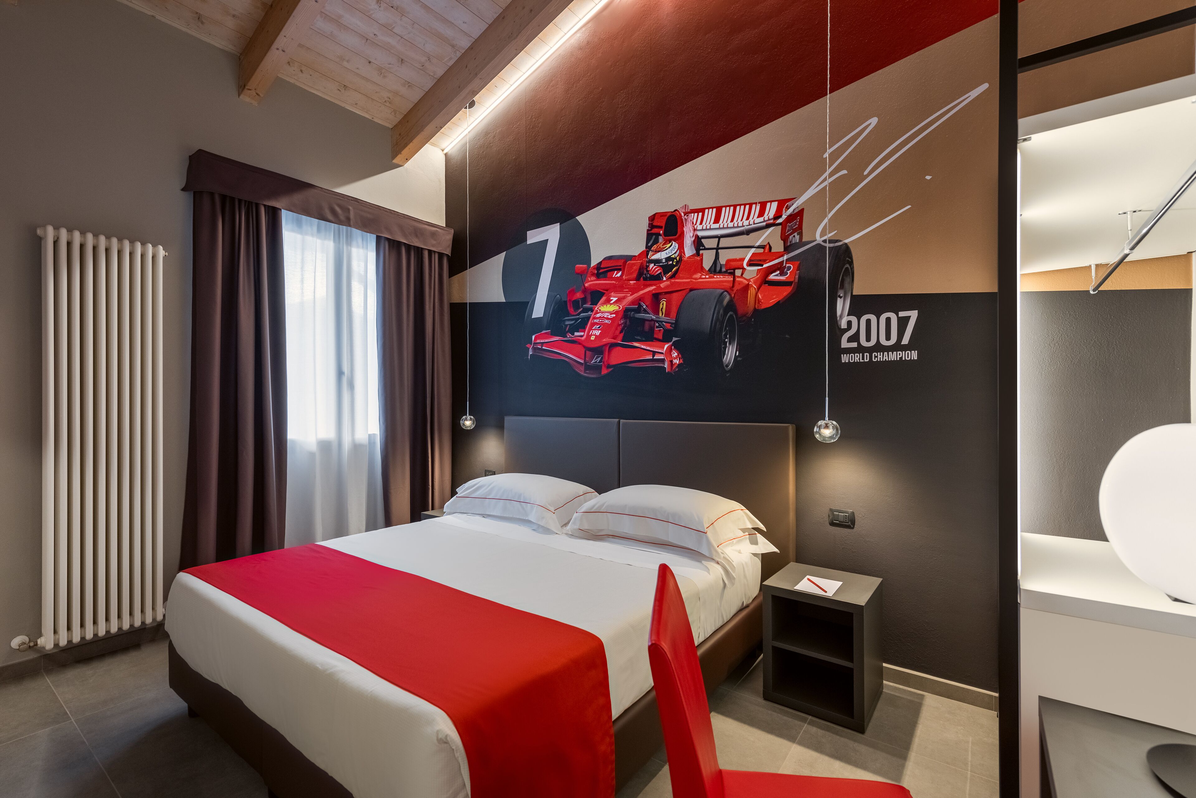 Una habitación de hotel moderna con una cama grande, un sillón rojo de acento, un mural de un auto de carreras y un ventanal con cortinas.