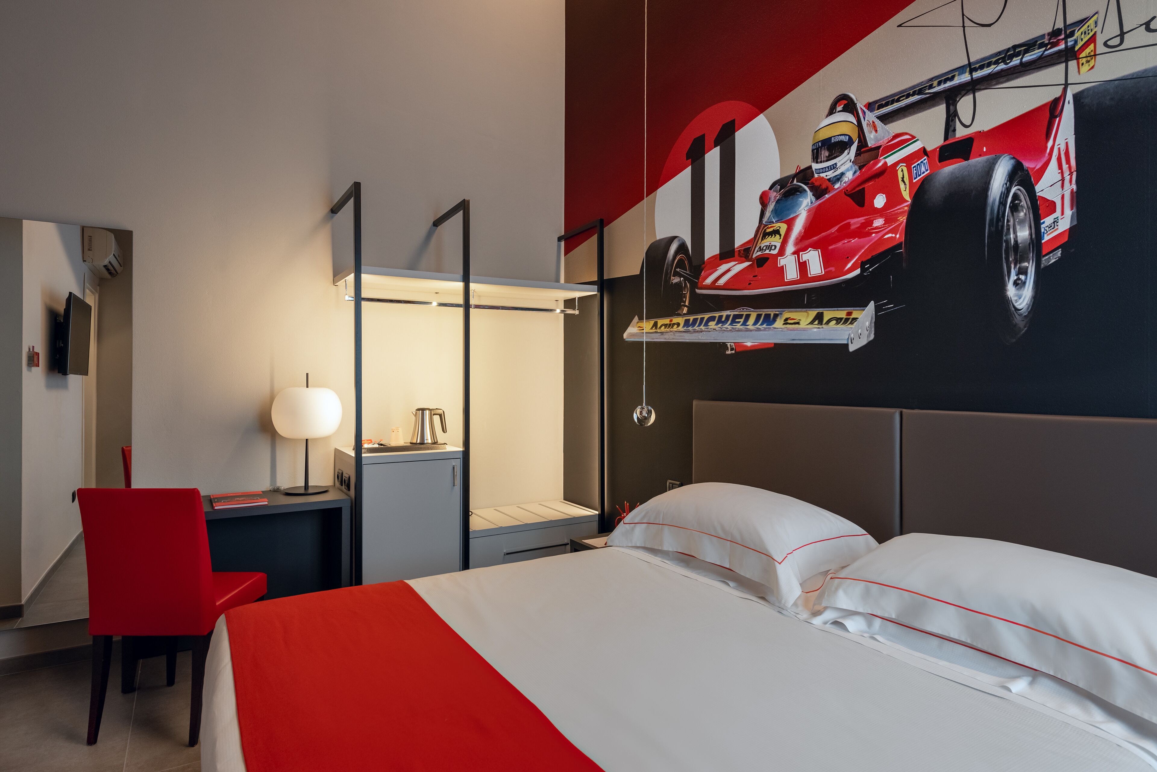 Een hotelkamer met een bed, een bureau met een lamp, een stoel en een groot wanddecor van een raceauto.