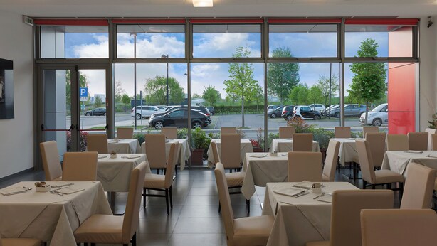 A restaurant with tables and chairs, large windows, and a view of parked cars outside. - Hotel Maranello Village (Maranello)