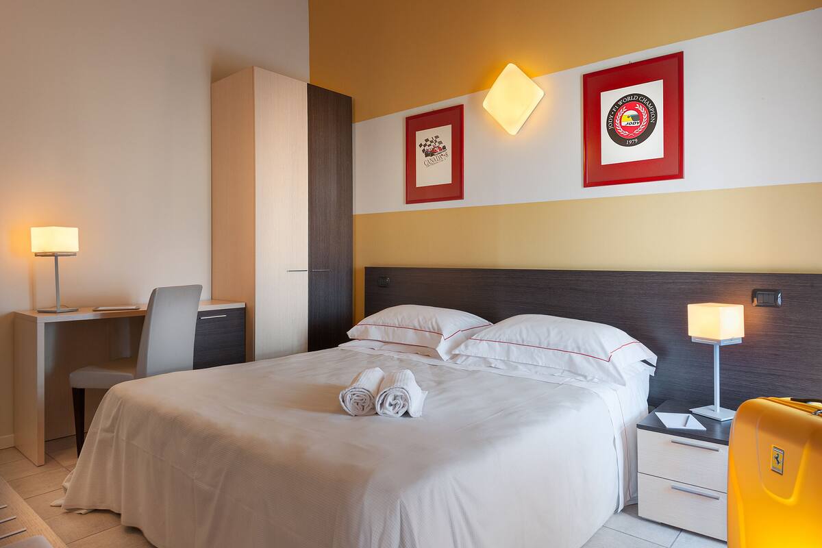 A neatly made bed with white linens and two rolled towels, a desk with a chair, a bedside table with a lamp, and two framed awards on the wall.