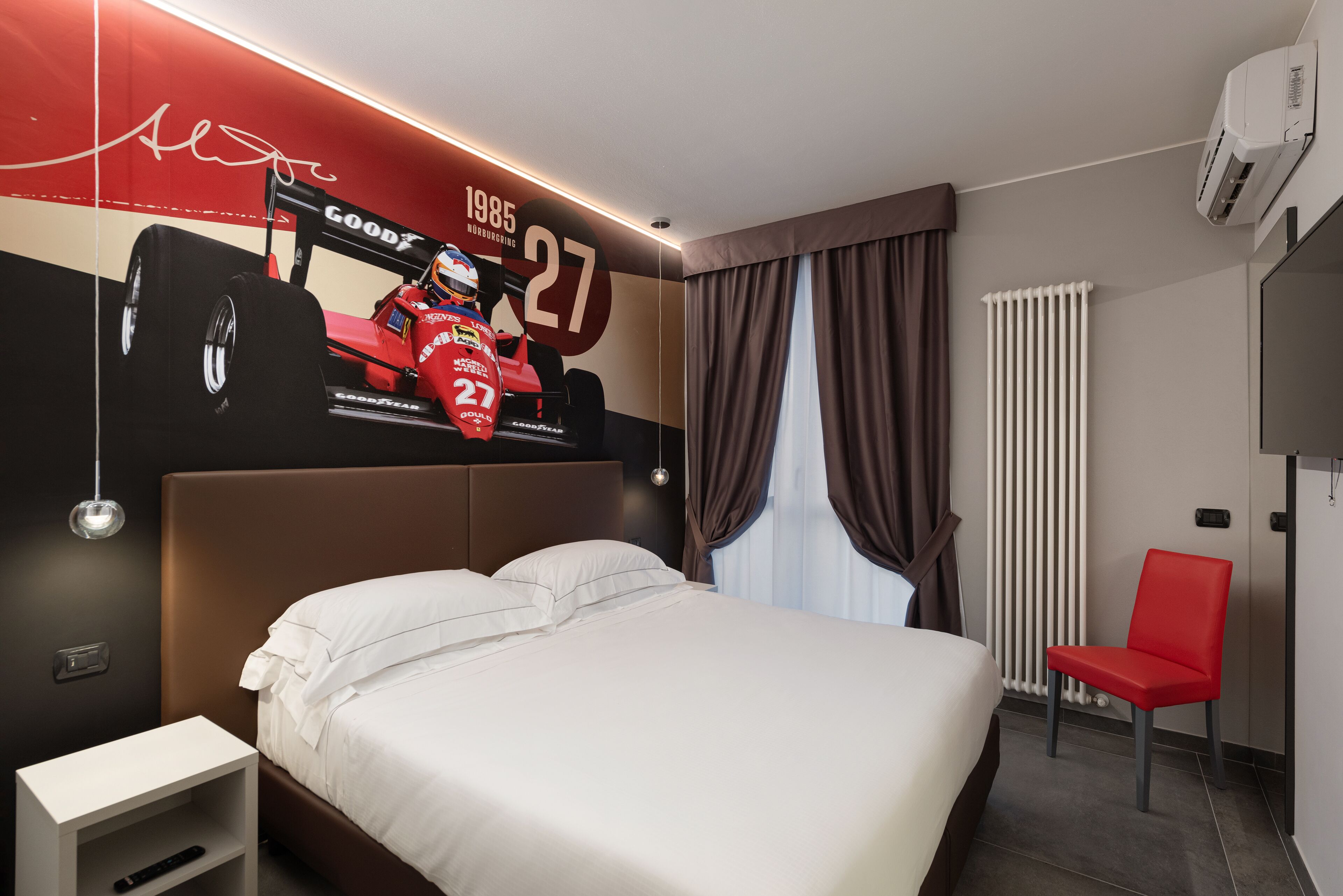 Une chambre d’hôtel moderne avec un grand lit, une affiche d’une voiture de course rouge accrochée au mur, un fauteuil rouge et une fenêtre avec des rideaux.