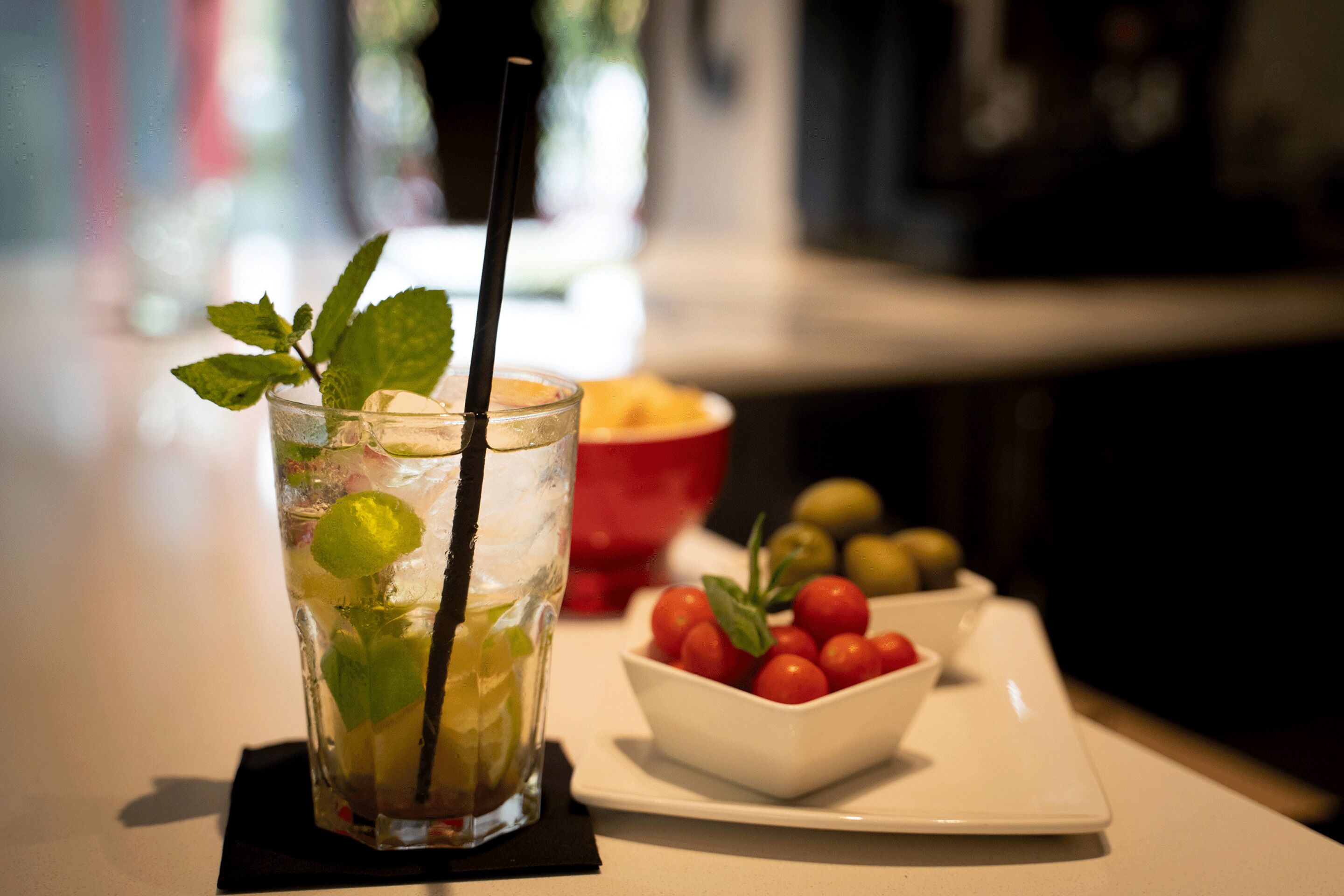 Un vaso de bebida helada con hojas de menta y una pajita, un cuenco de tomates cherry y un plato de aceitunas sobre una mesa.