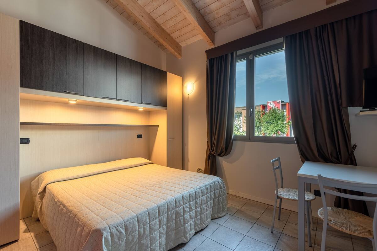 A bedroom with a bed, a desk, and a chair. There is a window with curtains, a ceiling with wooden beams, and a wall-mounted light fixture.