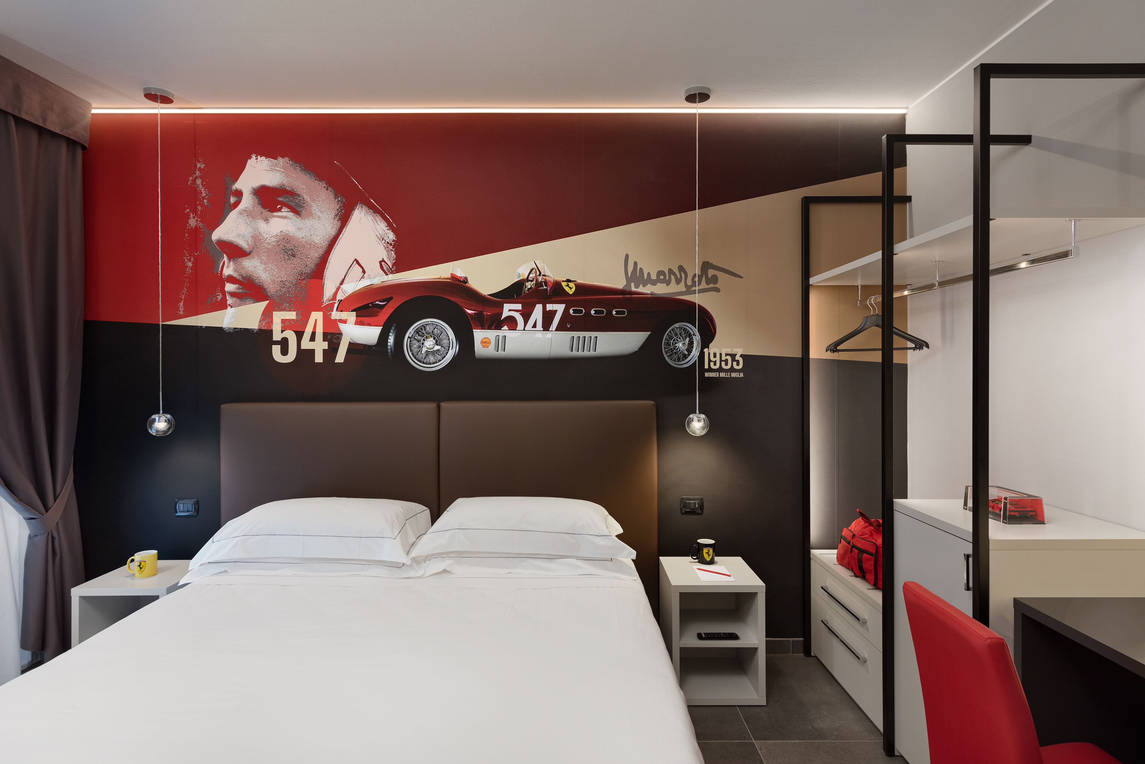 A modern hotel room with a large bed, a desk, a chair, a wardrobe, and a wall with a sports car mural.