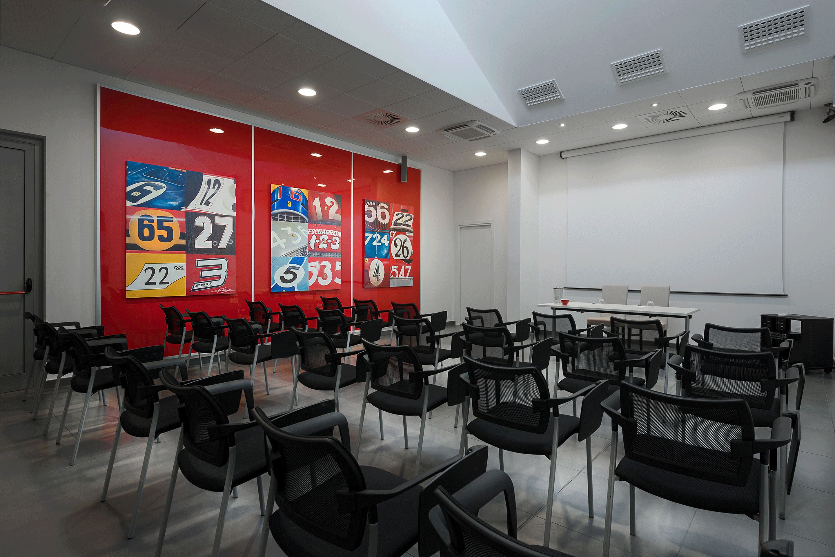 A conference room with rows of black chairs, a white table, and a red wall with numbered posters.