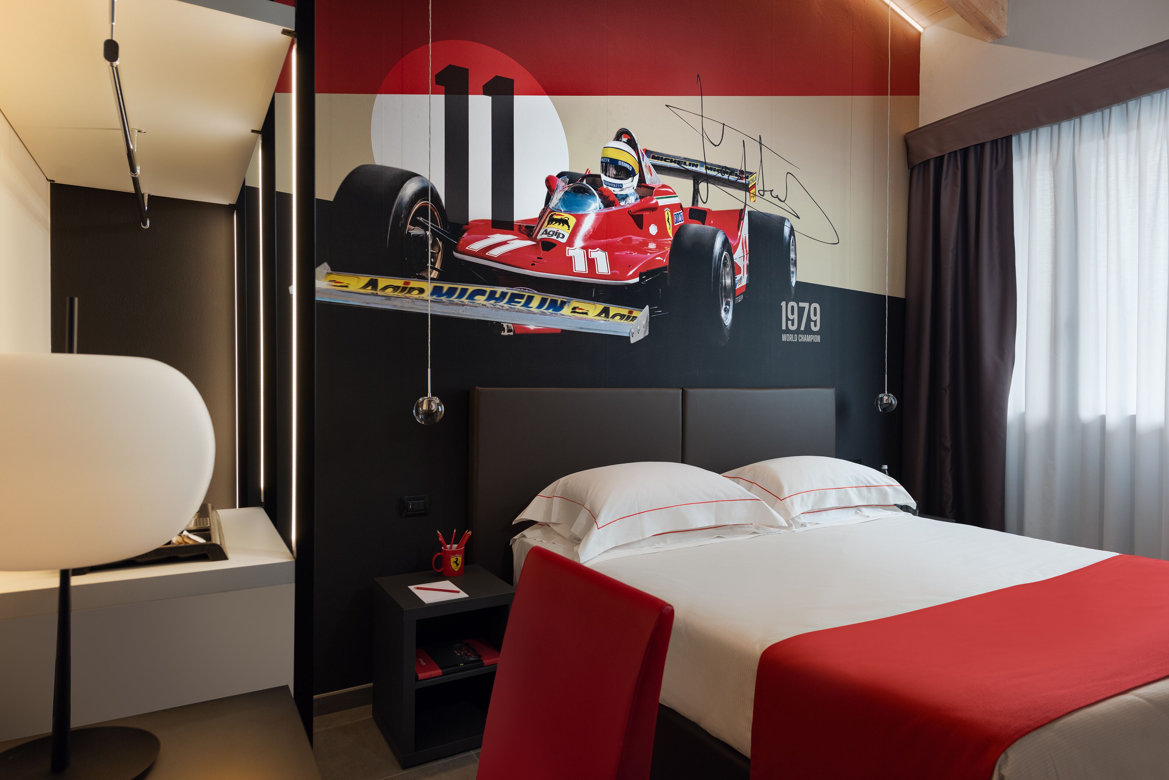 Camera d'albergo con un letto grande, una scrivania con una sedia, un comodino e un'opera d'arte appesa al muro raffigurante un'auto da corsa.
