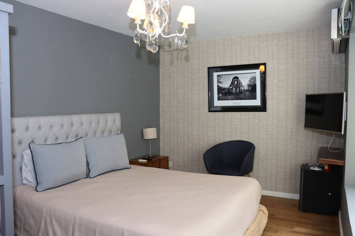 Ein Hotelzimmer mit einer gebügelten Kopfteil, einem Bett mit hellen Leinen, einem gerahmten Bild an der Wand, einem Fernseher auf einem Schrank und einem kleinen Tisch mit einer Lampe.