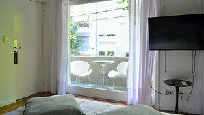 A hotel room with a bed, a TV, a balcony with chairs, and a view of buildings.