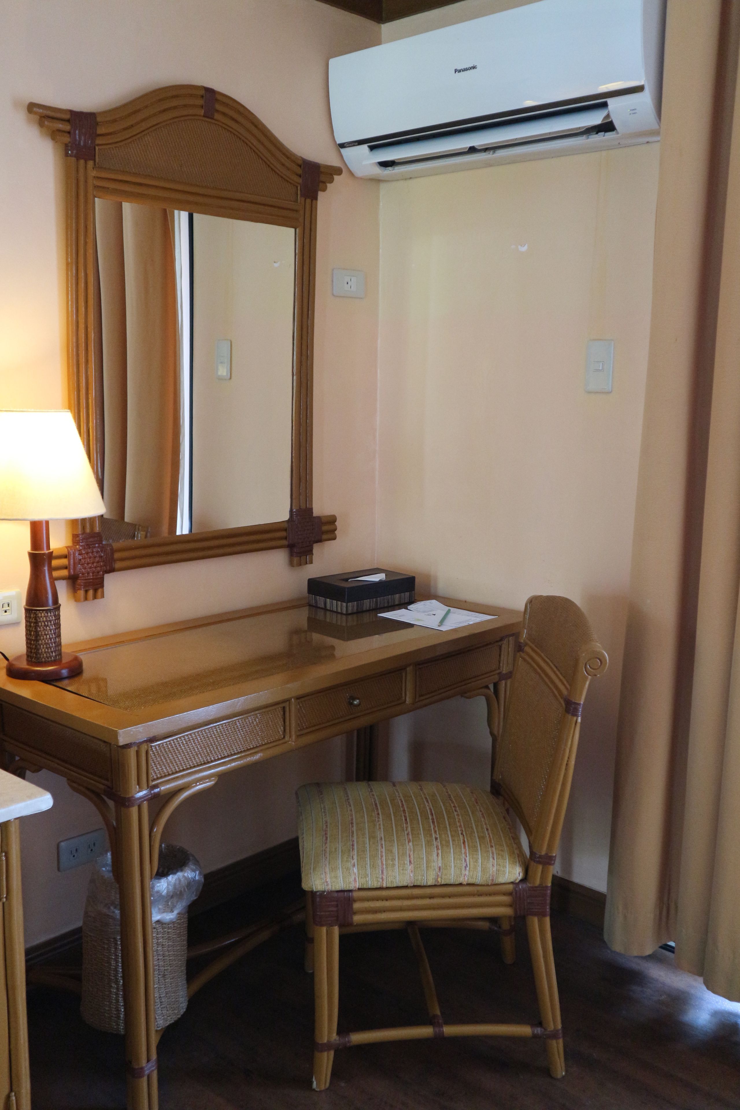 A hotel room with a wooden desk, a chair, a lamp, a mirror, and an air conditioning unit.