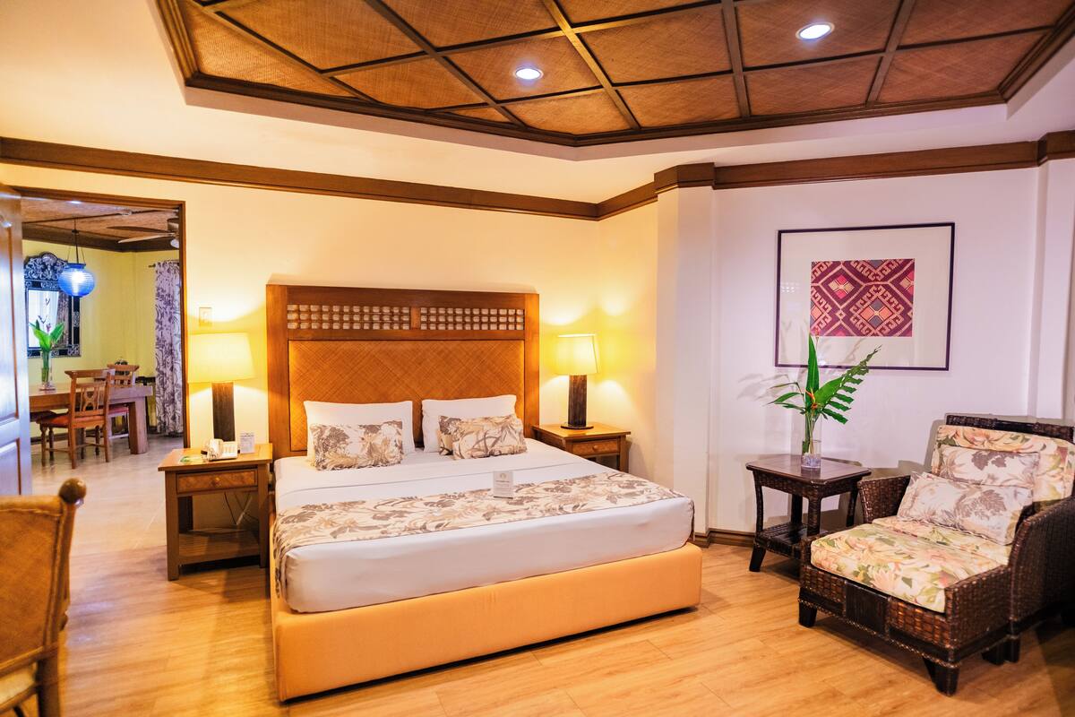 Habitación de hotel con una cama grande, dos mesitas de noche, una silla y un área de comedor visible a través de una puerta abierta.