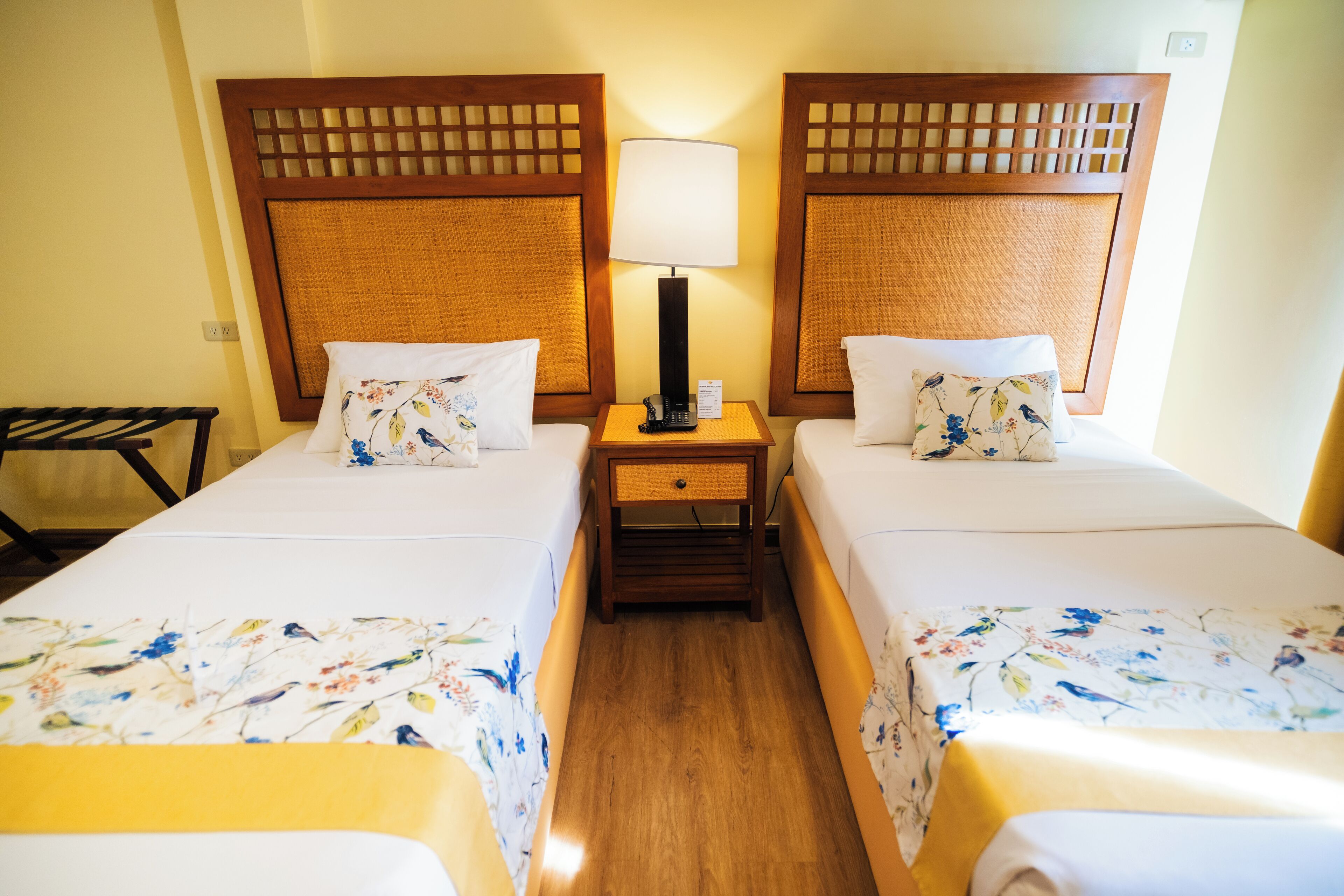 A hotel room with two single beds, each with a floral bedspread and matching pillows. There is a wooden headboard with a lattice design, a bedside table with a lamp, and a small drawer.