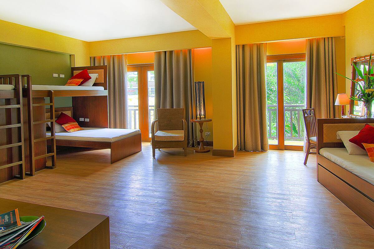 Habitación de hotel con una cama litera, una cama individual, una silla, una mesa y vistas a la vegetación a través de las ventanas.