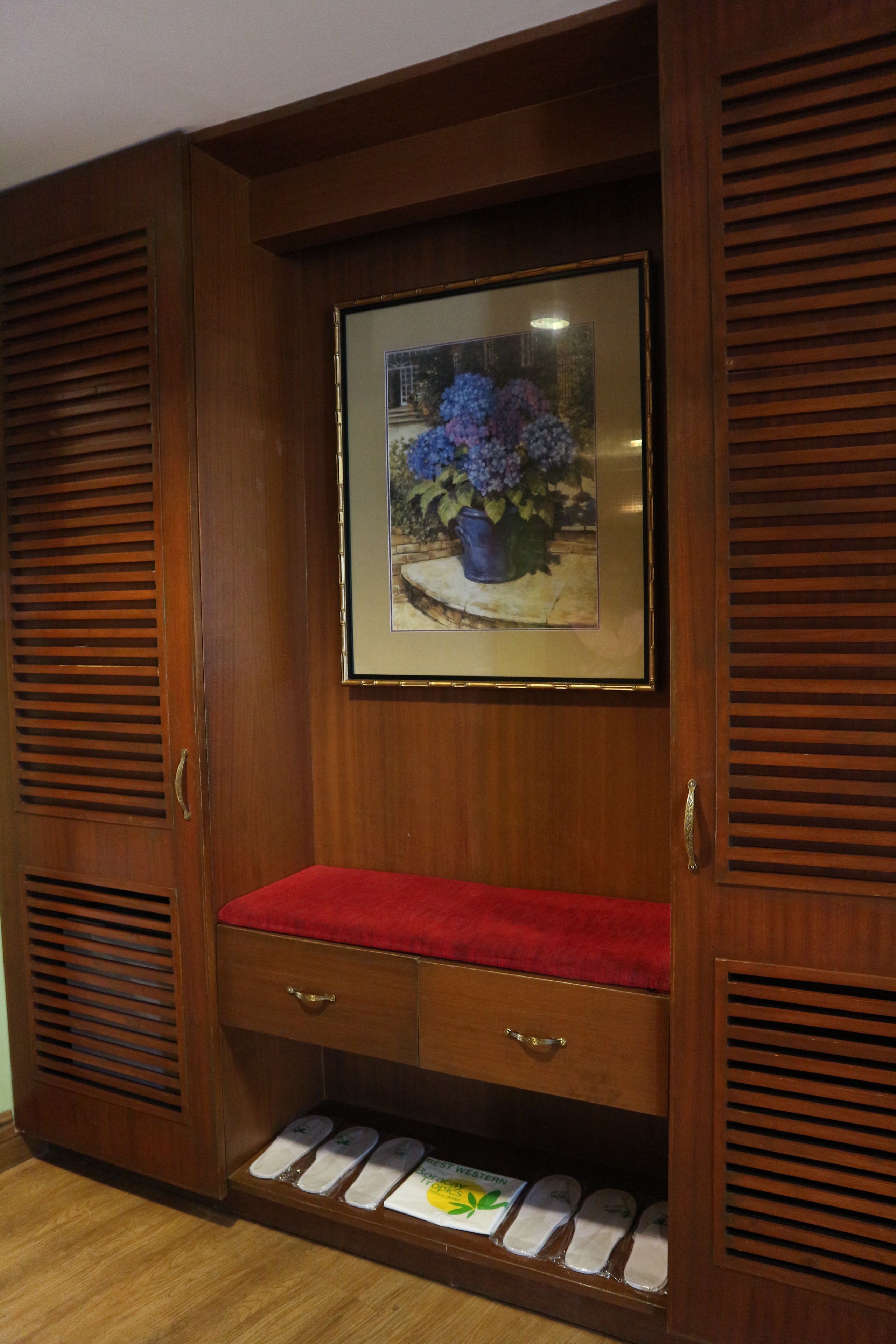 Een houten garderobe met een rode, beklede bank, een lijst met bloemen en een plank met pantoffels.