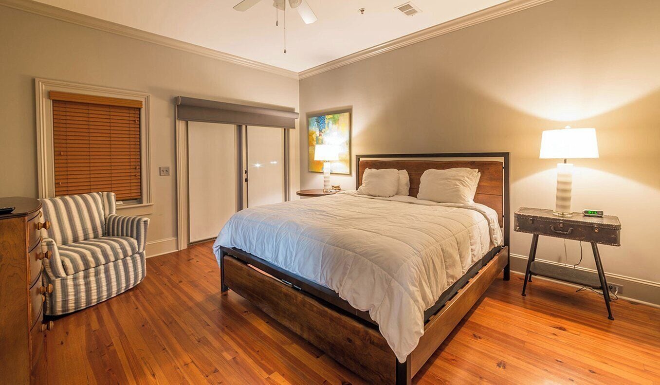 A bedroom with a wooden bed, a chair, a bedside table with a lamp, and a painting on the wall.