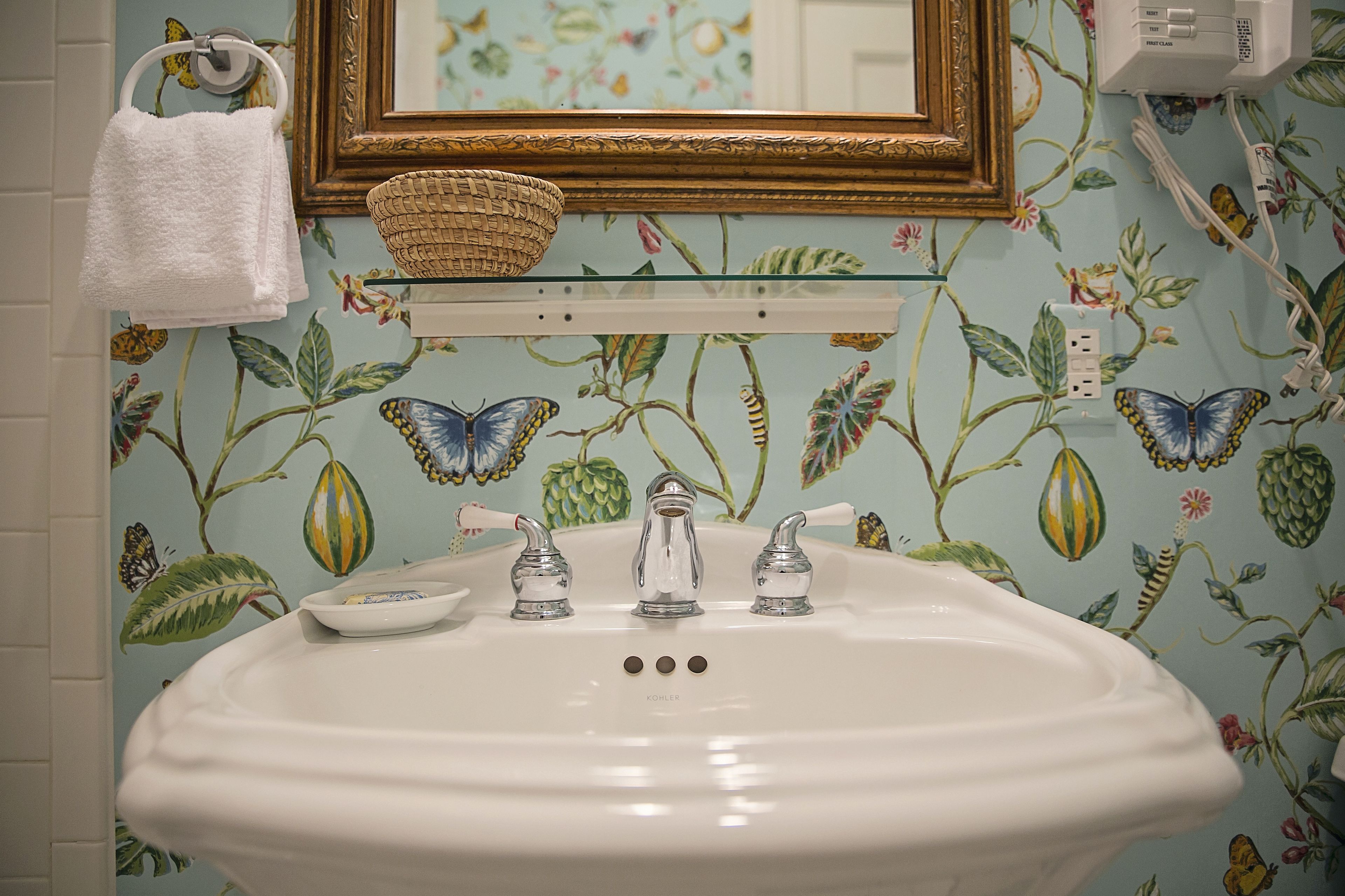 Een badkamer met bloemenbehang, een witte wastafel en een spiegel met gouden lijst.