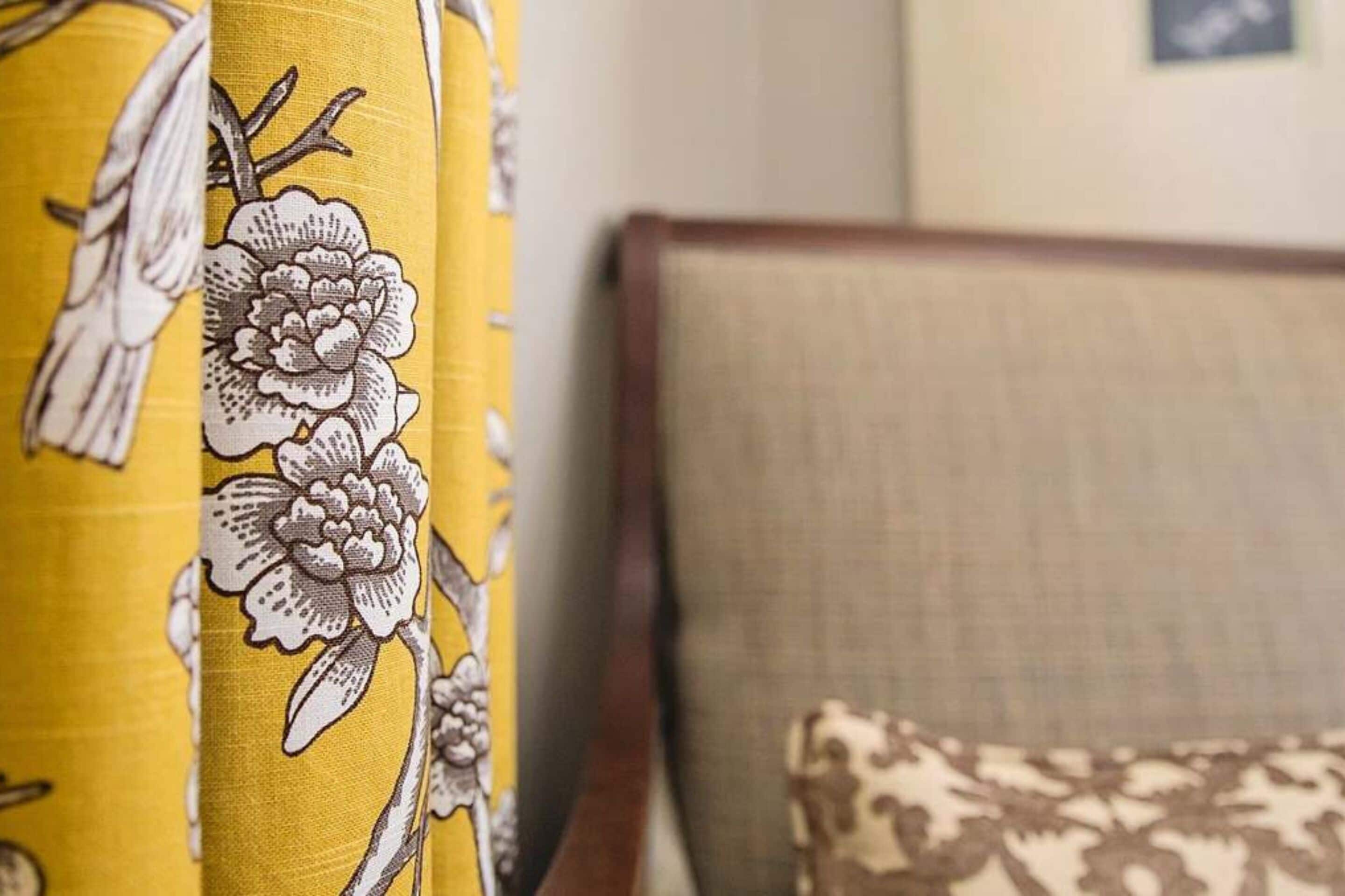 A close-up of a yellow curtain with a floral pattern, a beige upholstered headboard, and a patterned pillow.