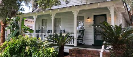 A white house with a porch, brick steps, and lush greenery.