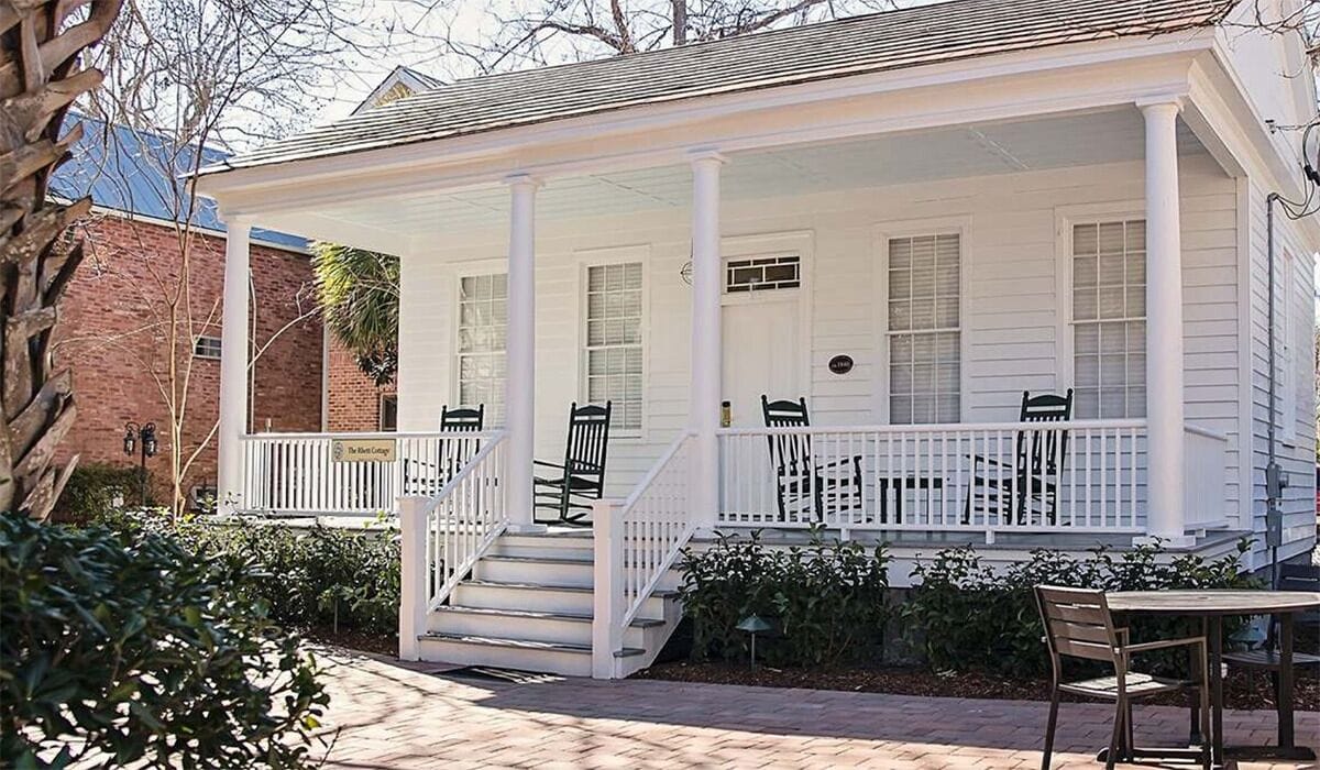 A white porch with a covered staircase, outdoor seating, and a brick building in the background.