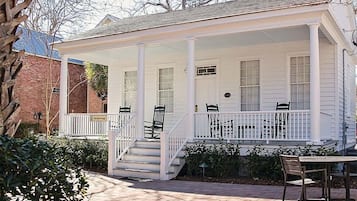 A white porch with a covered staircase, outdoor seating, and a brick building in the background.
