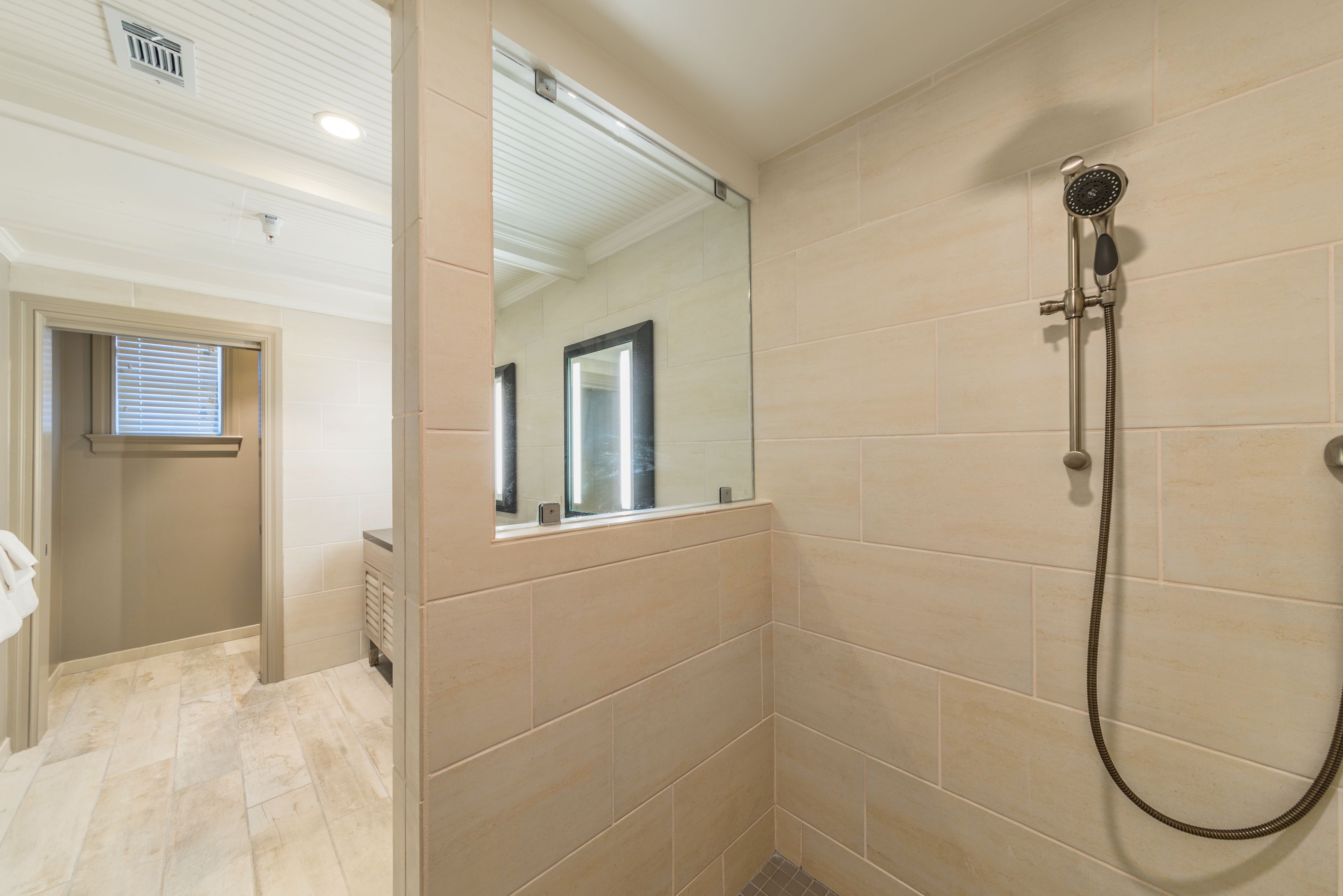 A bathroom with beige tiles, a shower with a handheld showerhead, and a mirror.