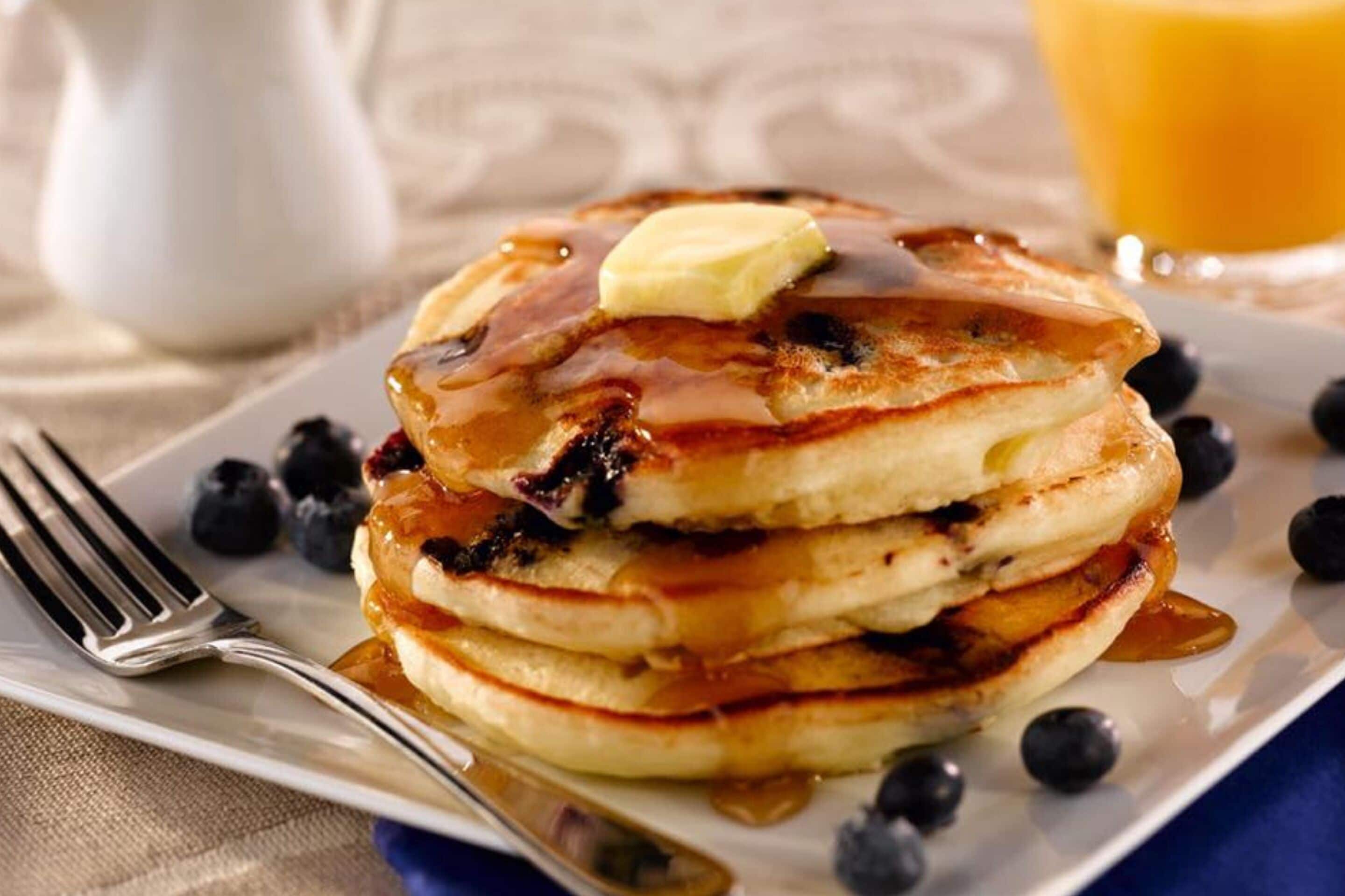 Una pila de tortitas con almíbar y un trozo de mantequilla encima, servidas en un plato blanco rodeado de arándanos.