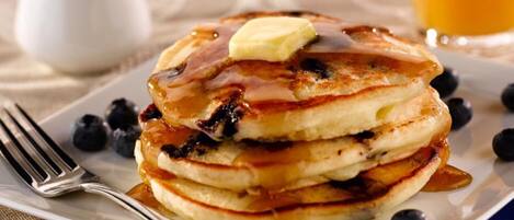 Una pila de tortitas con almíbar y un trozo de mantequilla encima, servidas en un plato blanco rodeado de arándanos.