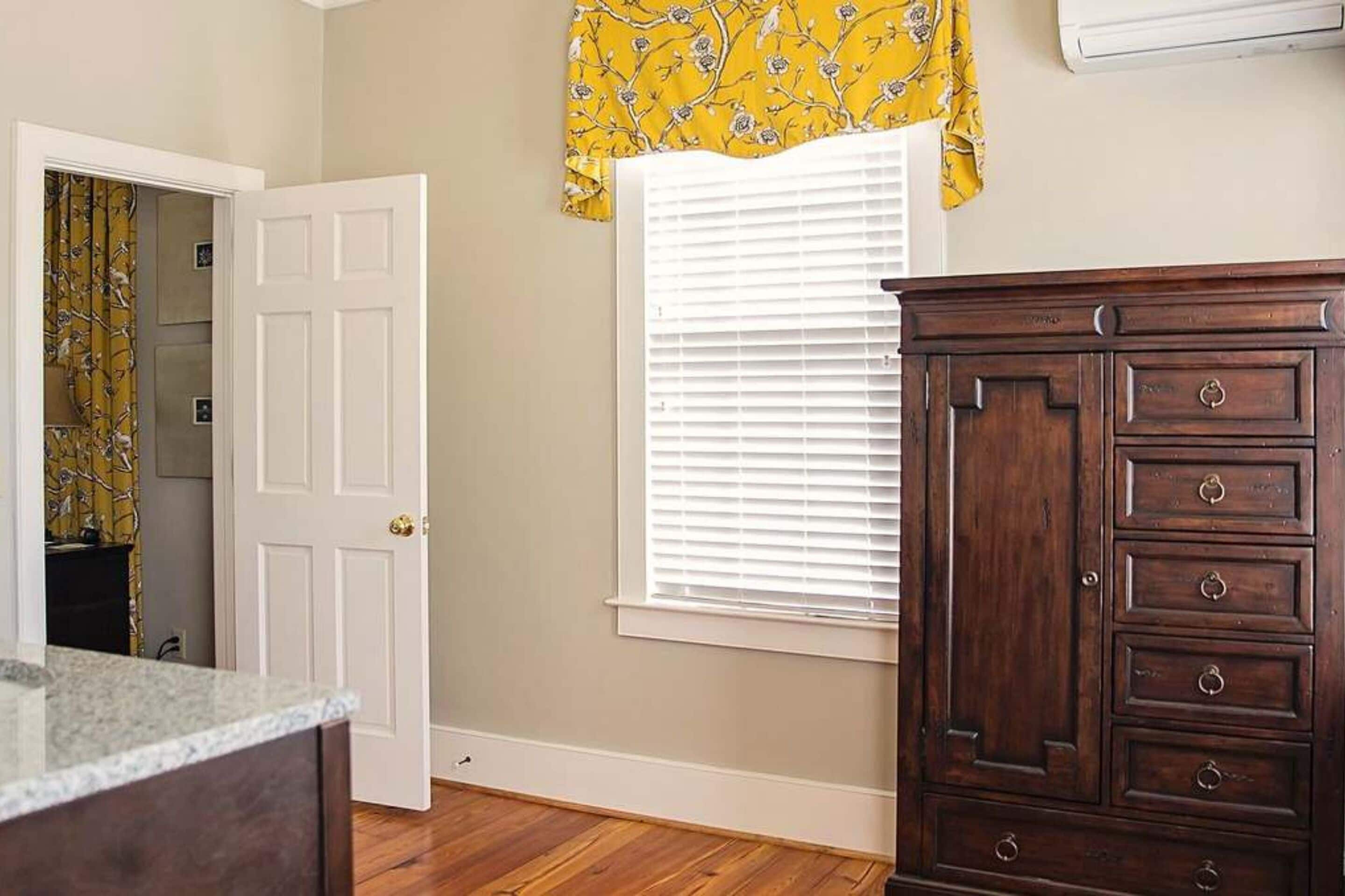 Une pièce avec un plancher en bois, une porte blanche, une fenêtre avec des stores et une commode en bois.