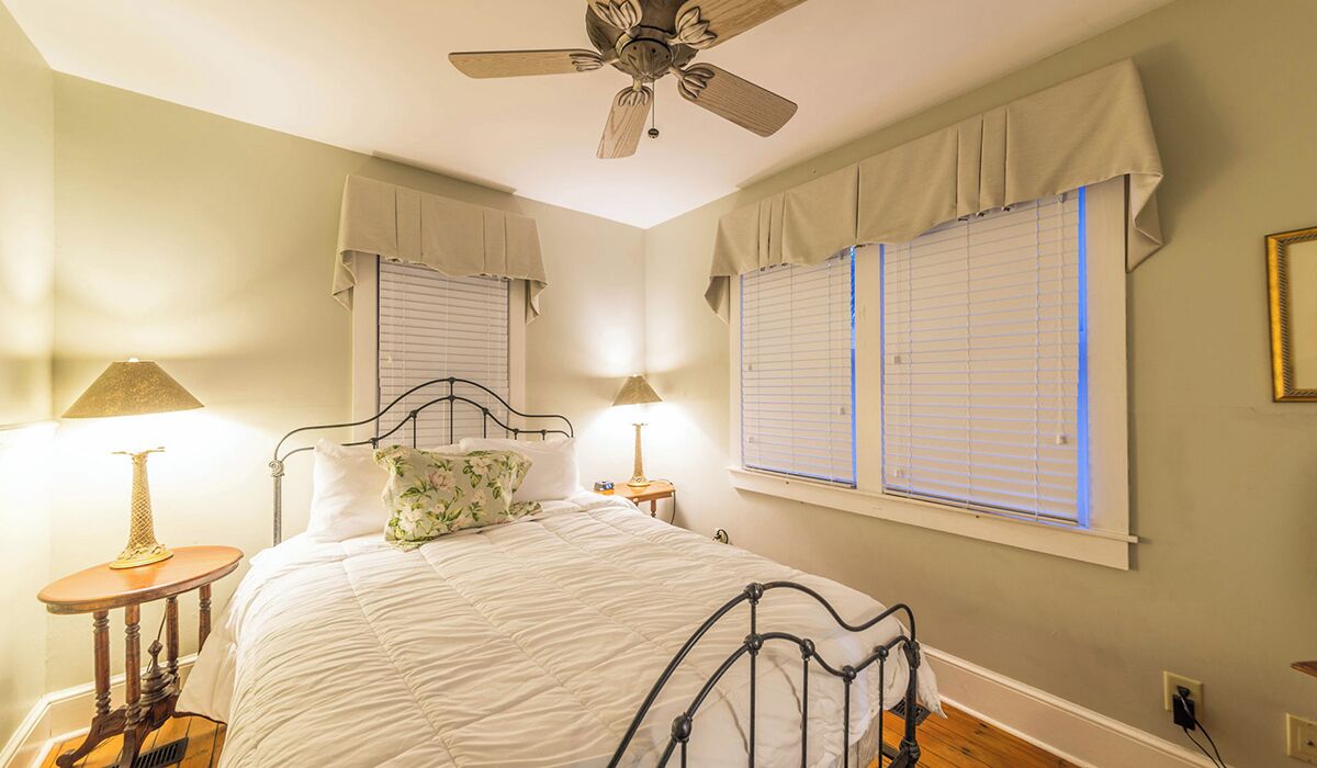 Une chambre à coucher comprenant un lit, deux tables de chevet, un ventilateur de plafond et des fen êtres équipées de stores.