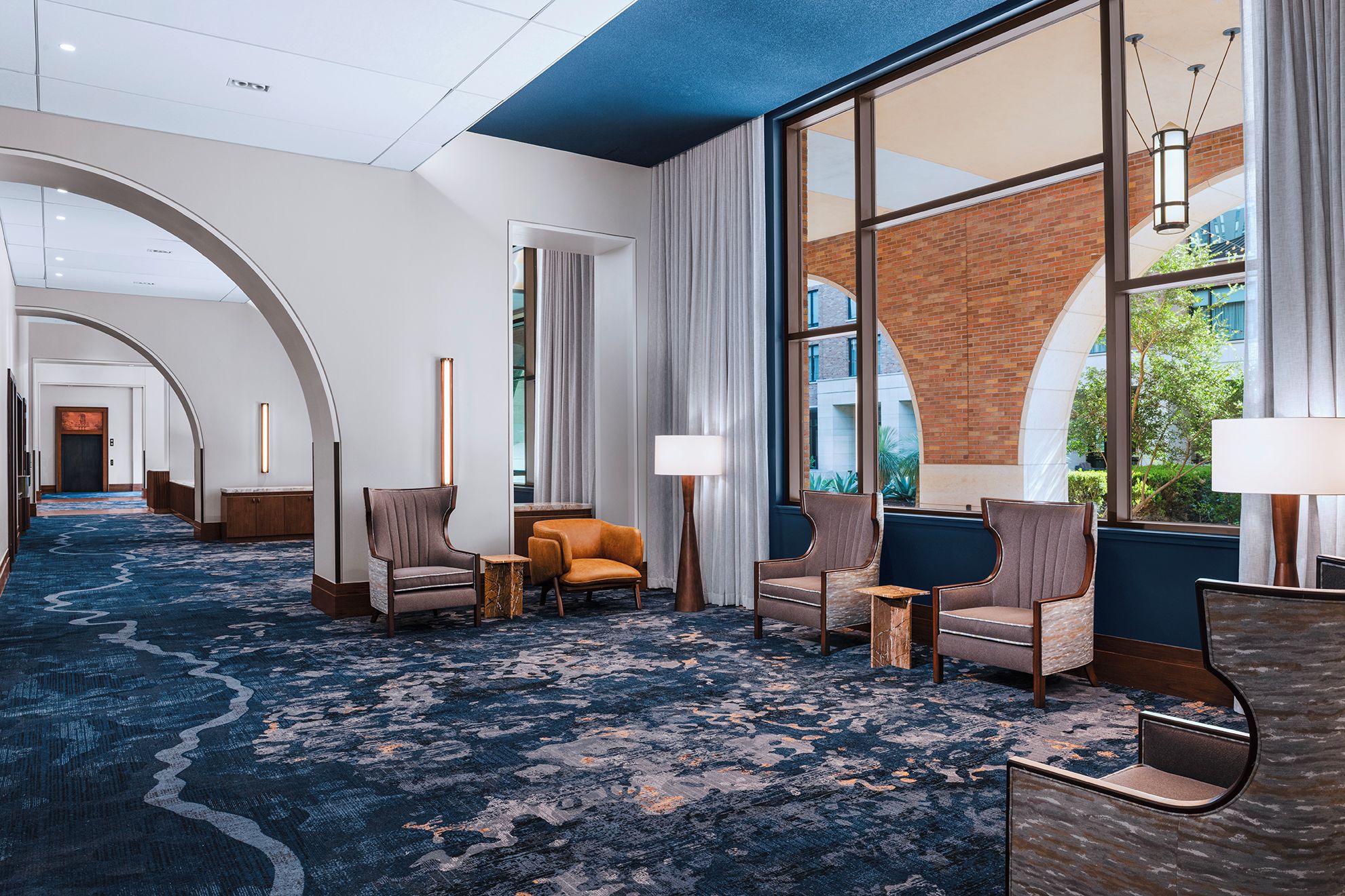 A hotel lobby with a blue carpet, seating area with chairs, a reception desk, and large windows offering views of the outside.