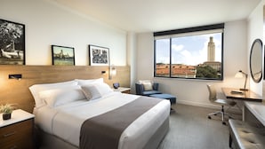 A hotel room with a large bed, a desk, a chair, and a view of a cityscape through a window. - AT&T Hotel & Conference Center at the University of Texas (Austin)
