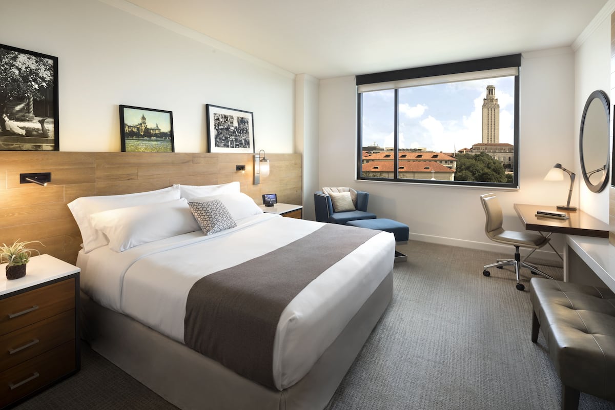 Habitación de hotel con una cama grande, un escritorio, una silla y vistas a un paisaje urbano a través de una ventana.