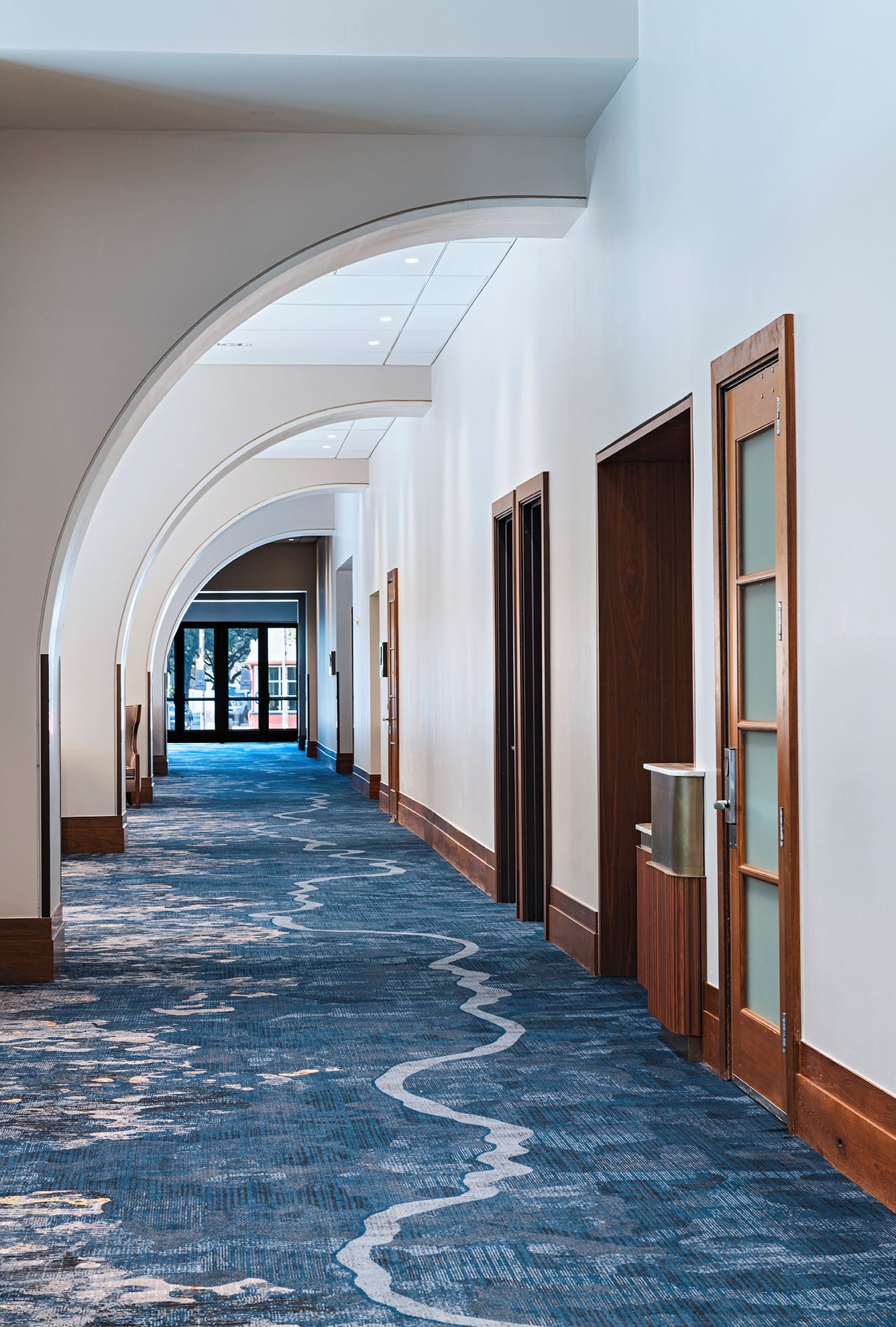 A hotel corridor with a blue carpet, wooden doors, and a skylight.